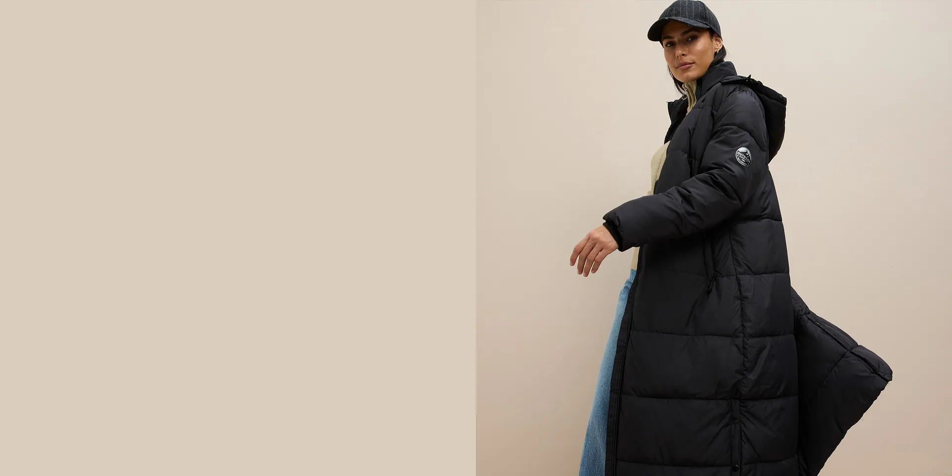 Doudoune longue noire portée sur un pull beige, un jean large et une casquette pour finaliser le look.