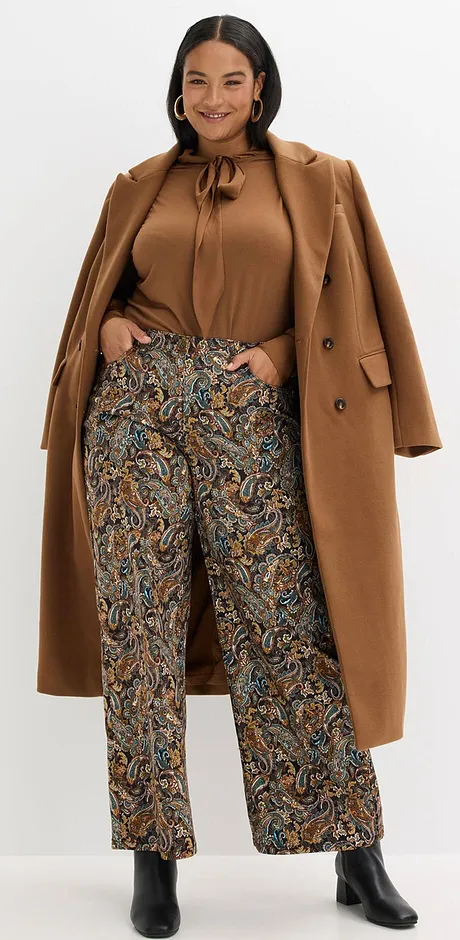 Manteau camel porté sur un pantalon à motif et une blouse à lavallière de couleur assortie.