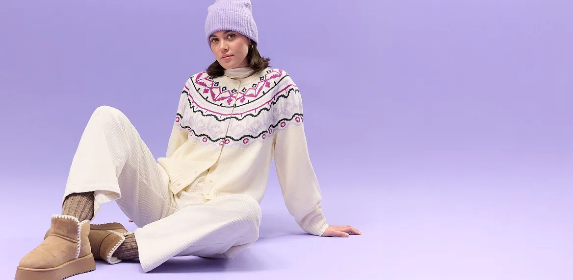 Cremefarbener Pullover mit nordischem Muster, weiße Hose und lila Strickmütze.