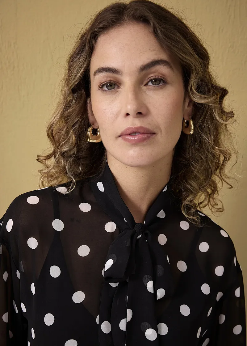 Elegante schwarze Bluse mit weißen Punkten und Schleifenkragen. Goldene Ohrringe vervollständigen den Look.