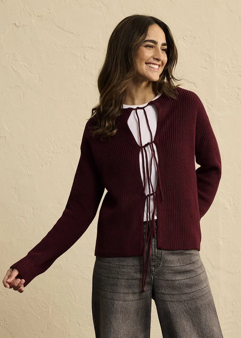 Kombination aus weinroter Strickjacke, weißer Bluse mit Schnüren und grauer Hose.