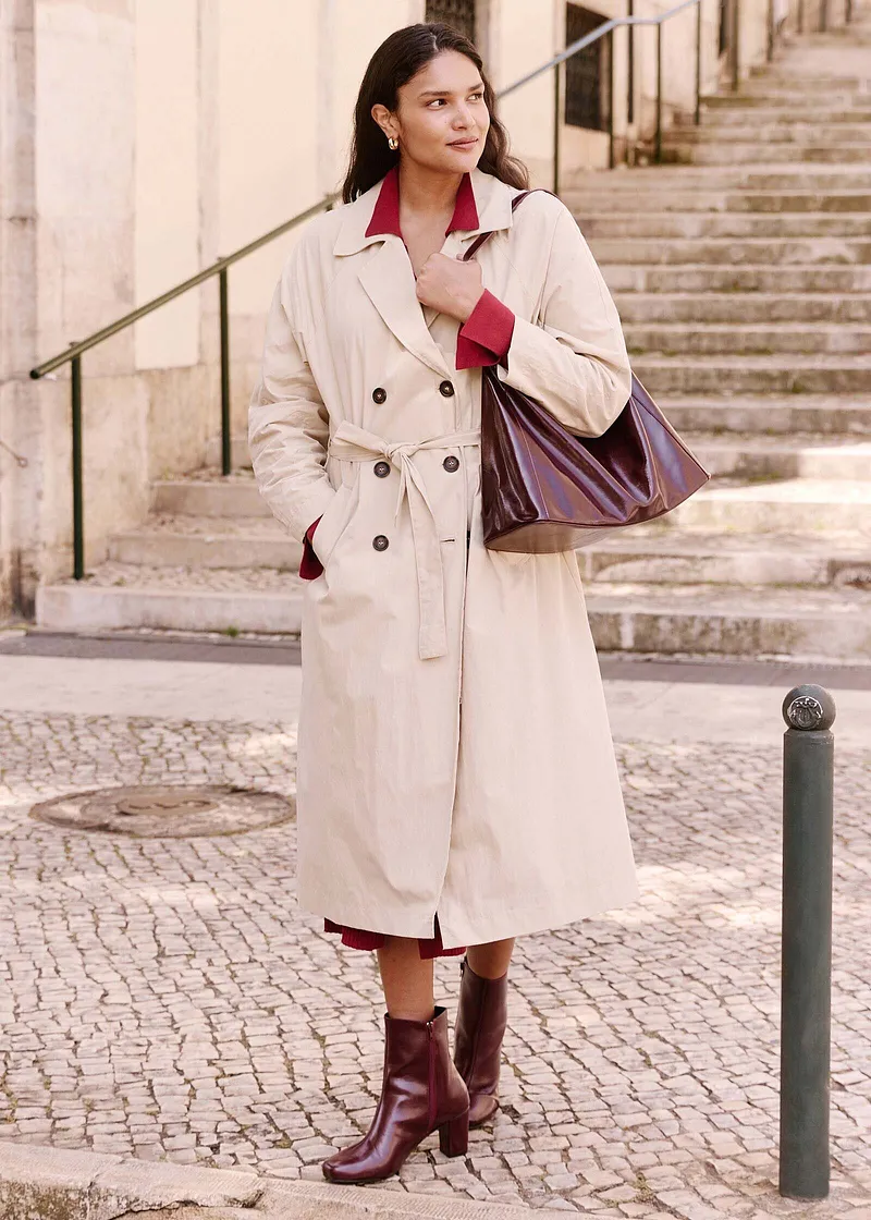 Eleganter cremefarbener Mantel mit Gürtel, rote Bluse und braune Stiefel. Stilvolle Handtasche vervollständigt den Look.
