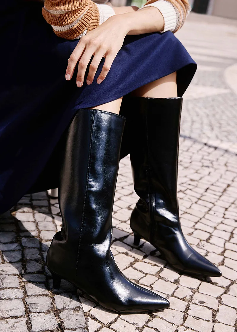 Eleganter Kontrast: Beige Pullover, dunkelblauer Rock und glänzende schwarze Lederstiefel auf Kopfsteinpflaster.