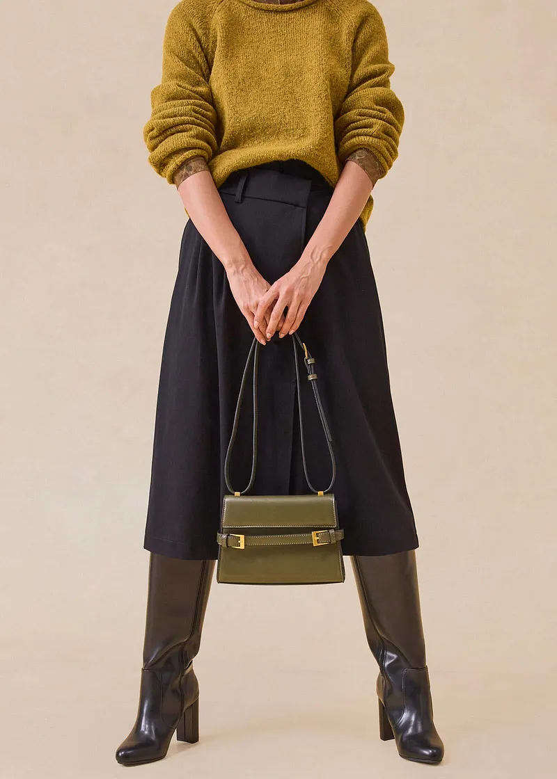 Pull jaune moutarde, jupe midi noire, bottes marron et sac à main élégant.