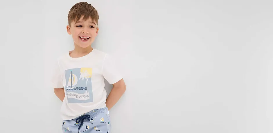 Enfant joyeux vêtu d'un T-shirt blanc avec un motif voilier et d'un pantalon bleu clair avec des motifs voiliers.
