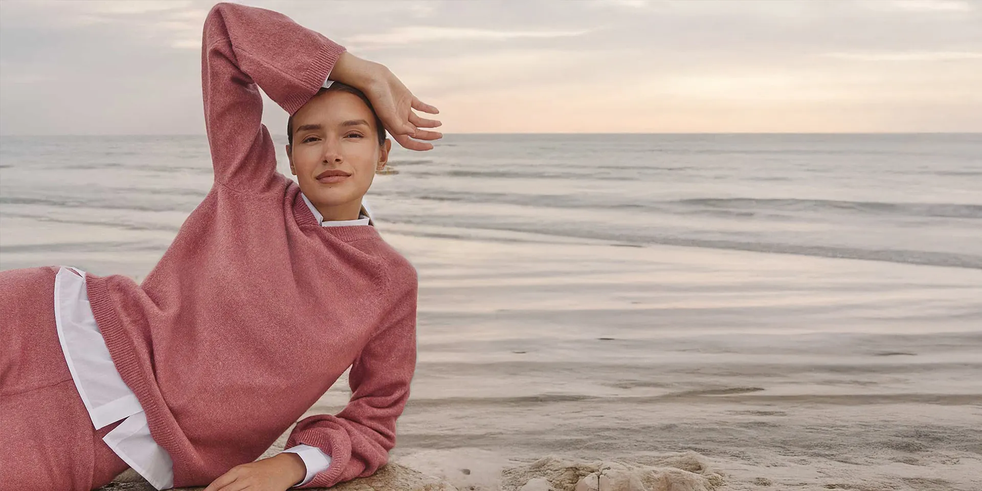 Frau in rosa Strick-Hoodie am Meer, gemütlicher Loungewear-Look.  