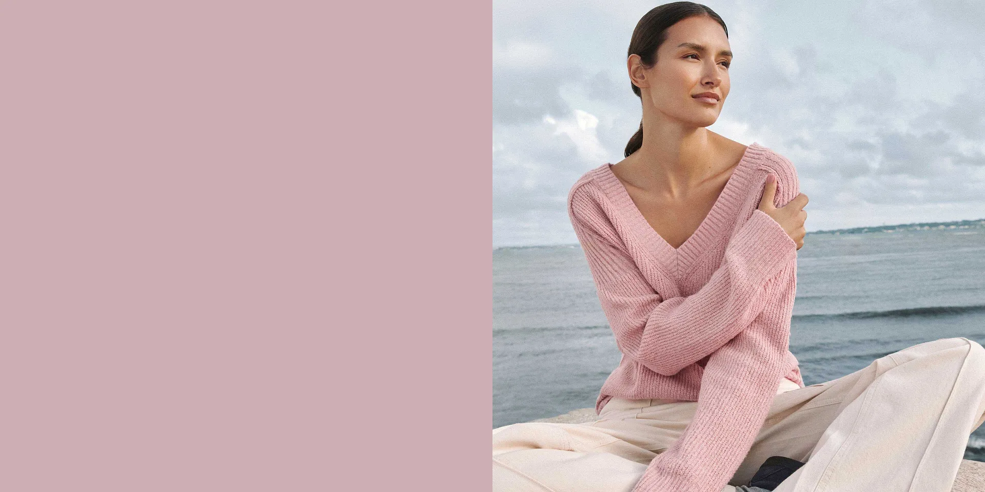 Ein zartrosa Strickpullover mit V-Ausschnitt schmückt die Gestalt vor dem Meereshintergrund.