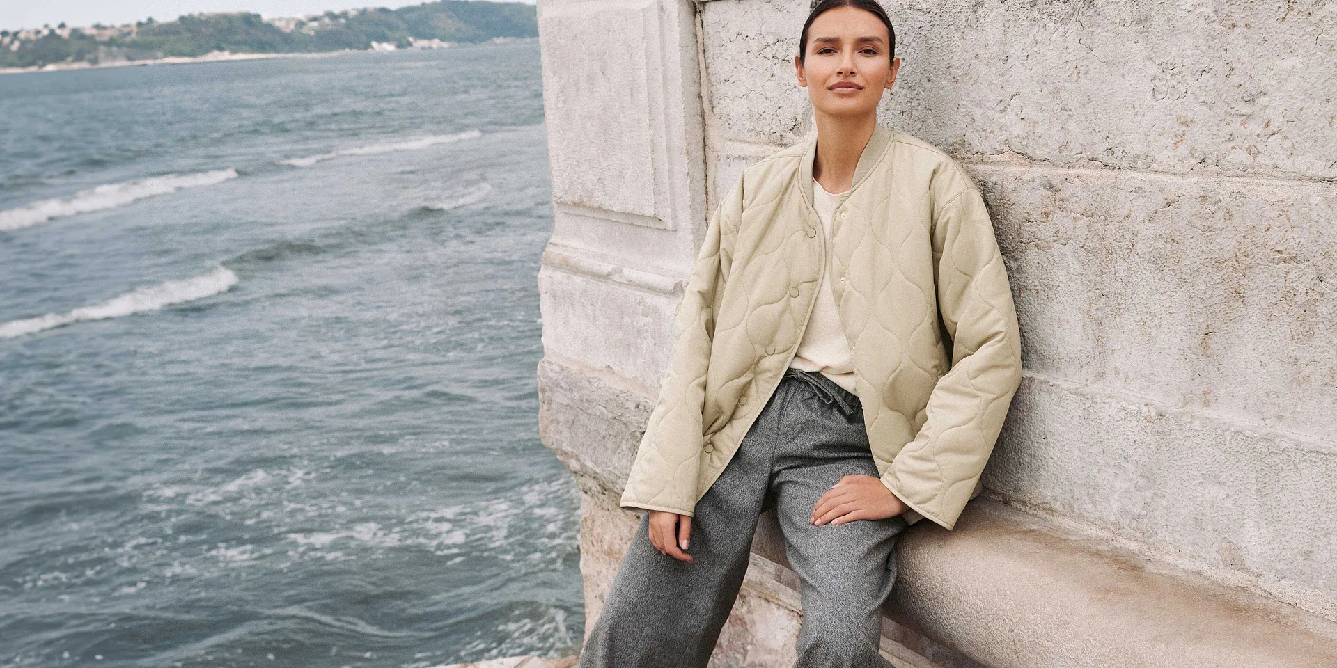 Lässiger Herbstlook: Beigefarbene Jacke, hellblaue Jeans und weißes Top am Wasser.