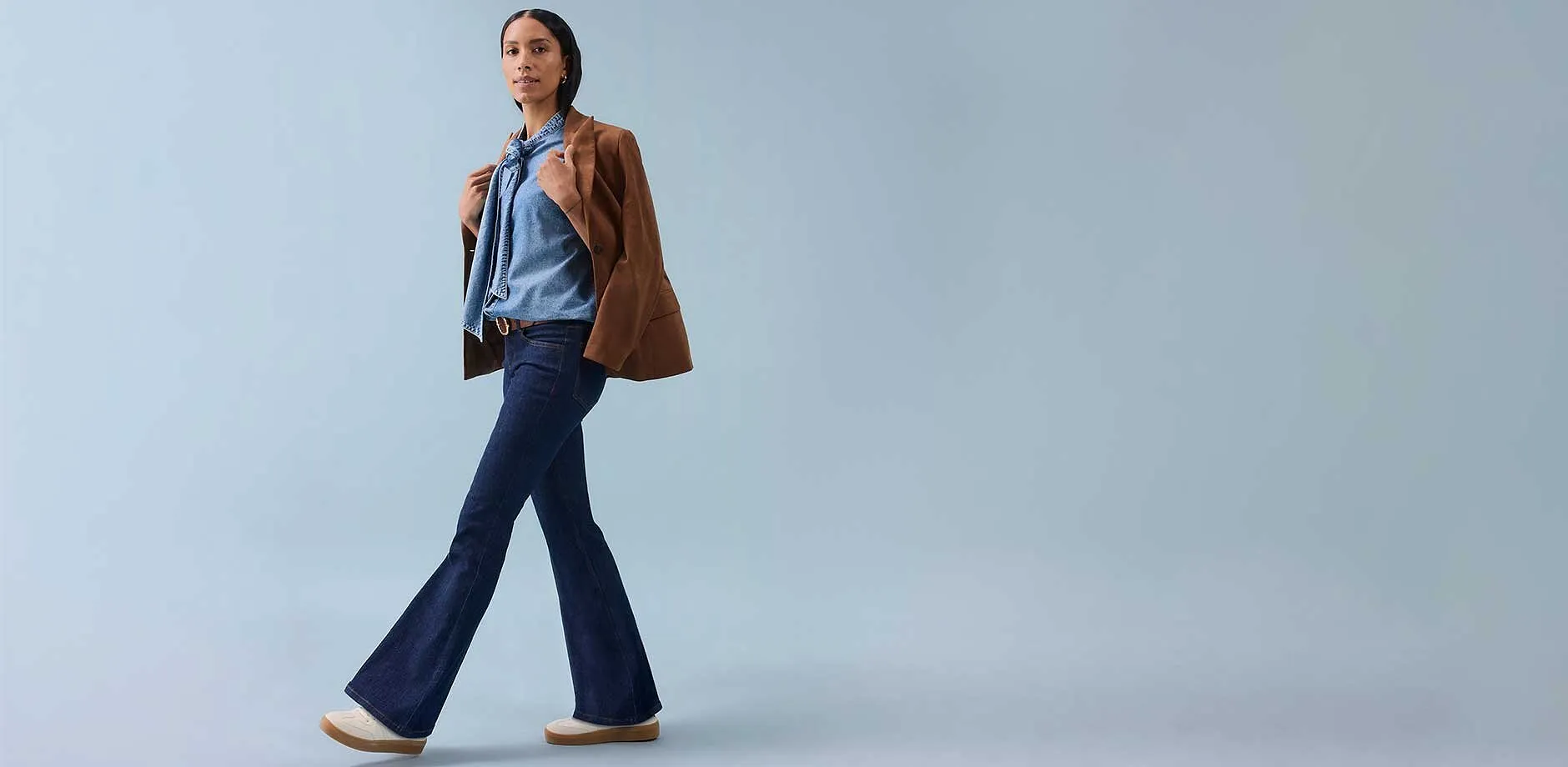 Eine Frau trägt eine Schlaghose, ein ärmelloses Top und eine braune Jacke lässig über der Schulter.