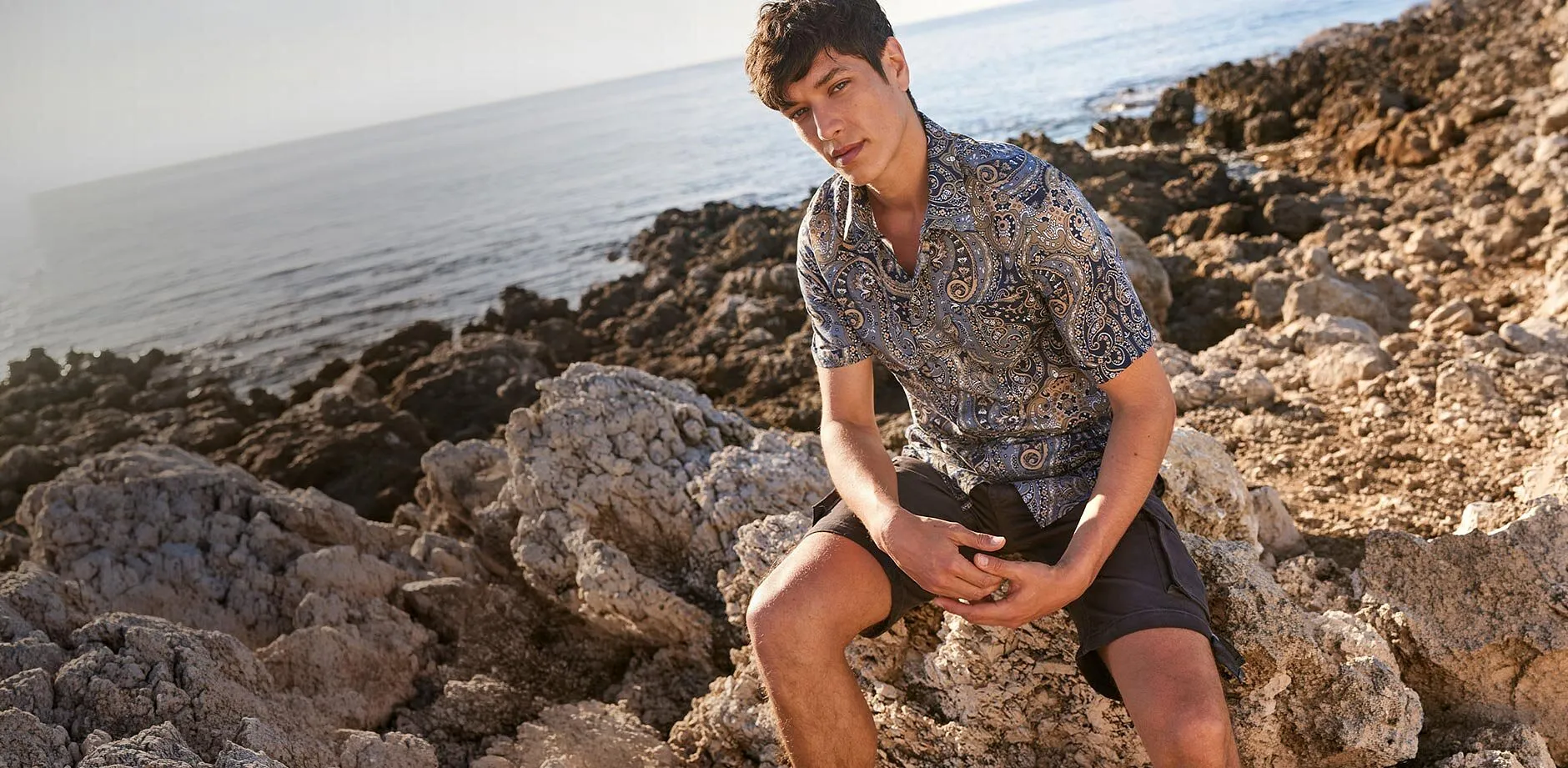 Un homme portant une chemise imprim&eacute;e et un short fonc&eacute; sur les rochers &agrave; la plage.