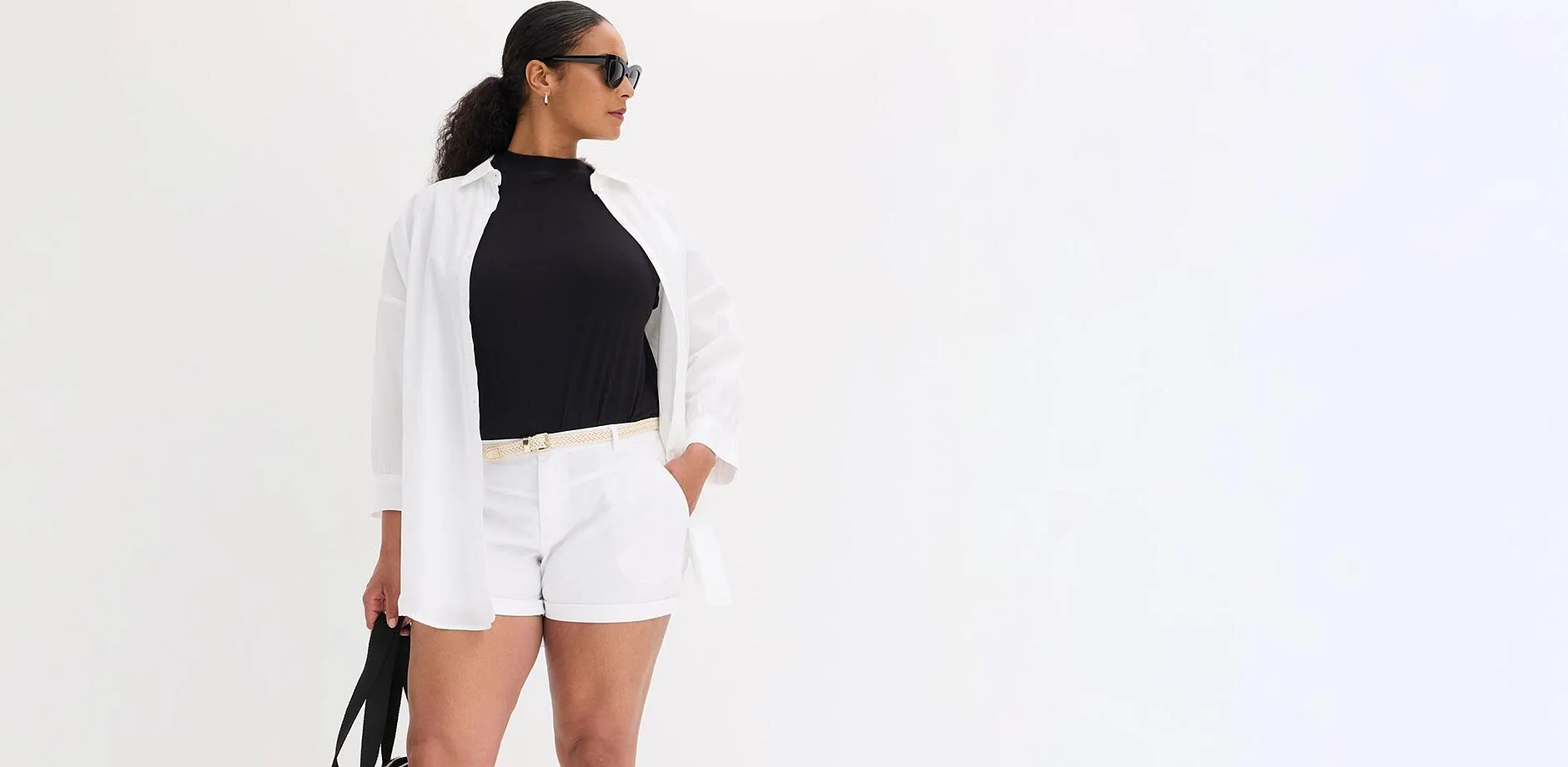 Chemise blanche ouverte sur un top noir, short blanc, ceinture claire, lunettes de soleil.