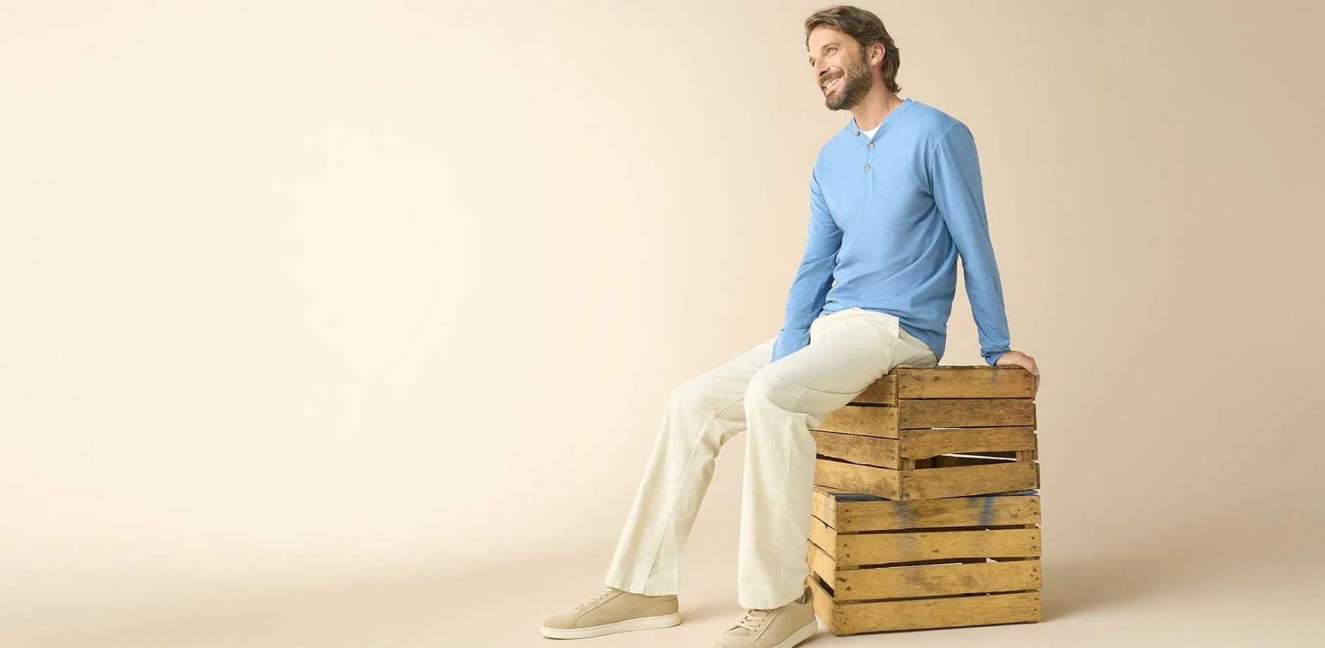 Er tr&auml;gt ein hellblaues Langarmshirt, eine helle Hose und beige Sneaker.