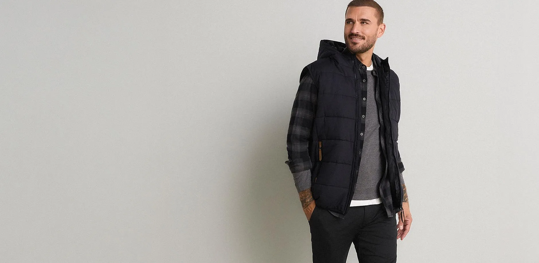 Gilet matelass&eacute; &agrave; capuche, chemise &agrave; carreaux, T-shirt gris et pantalon noir.