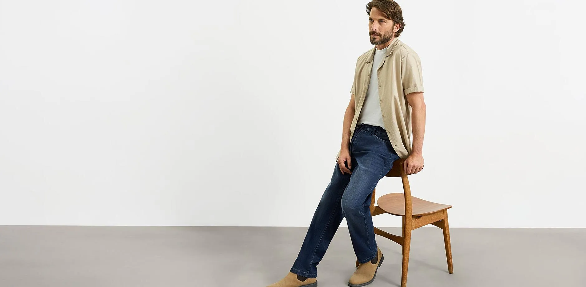 Beiges Hemd, graues T-Shirt, blaue Jeans und braune Wildlederschuhe bilden den Look.