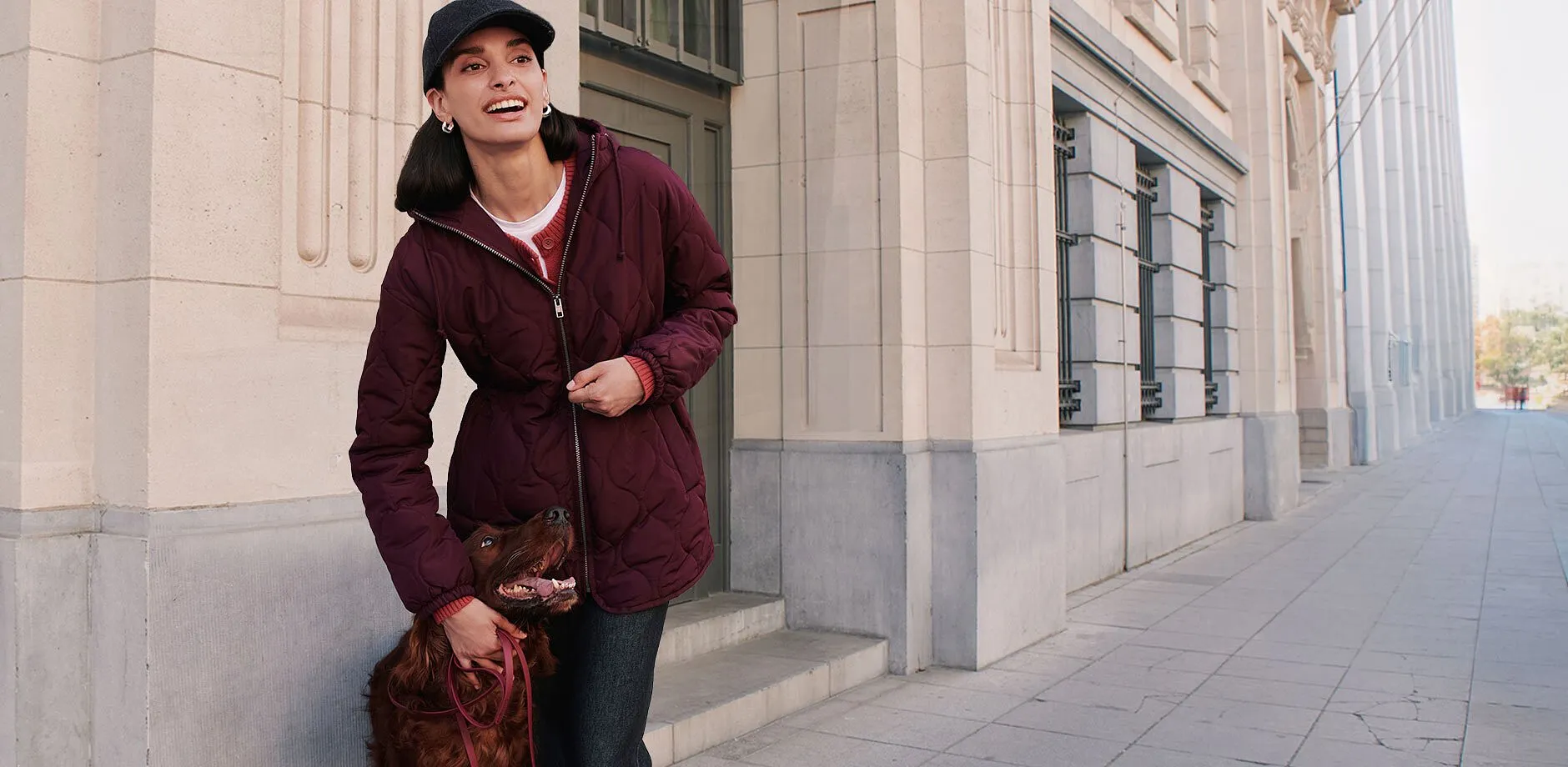 Dunkle Kappe, gesteppte bordeauxrote Jacke, roter Cardigan und dunkle Jeans.