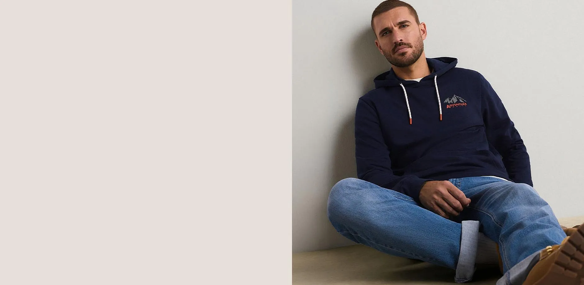 Er tr&auml;gt einen dunkelblauen Hoodie mit Bergmotiv und dazu eine l&auml;ssige blaue Jeans.