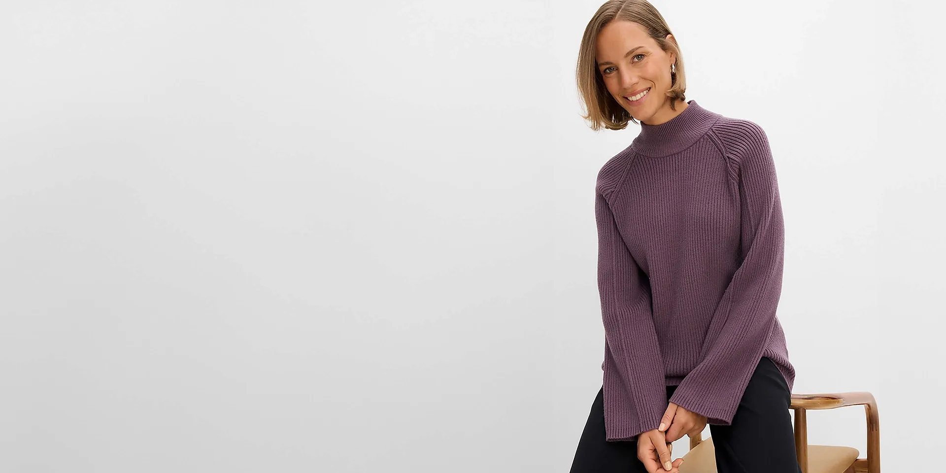Sie tr&auml;gt einen stilvollen violetten Rollkragenpullover aus Rippstrick und eine schwarze Hose.