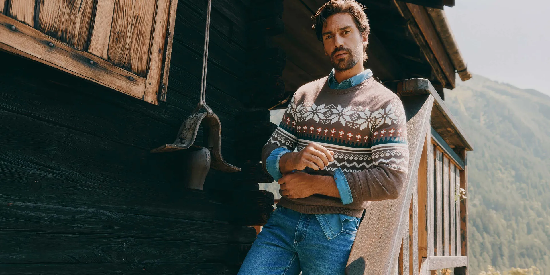 Er tr&auml;gt einen braunen Pullover mit Muster, ein Jeanshemd und blaue Jeans.