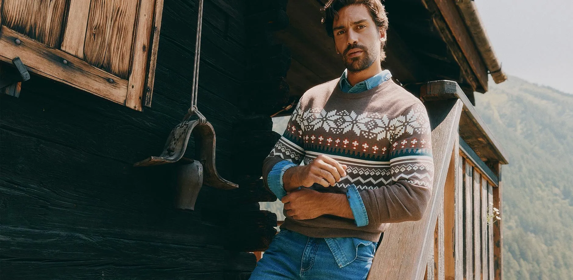 Er tr&auml;gt einen braunen Pullover mit Muster, ein Jeanshemd und blaue Jeans.