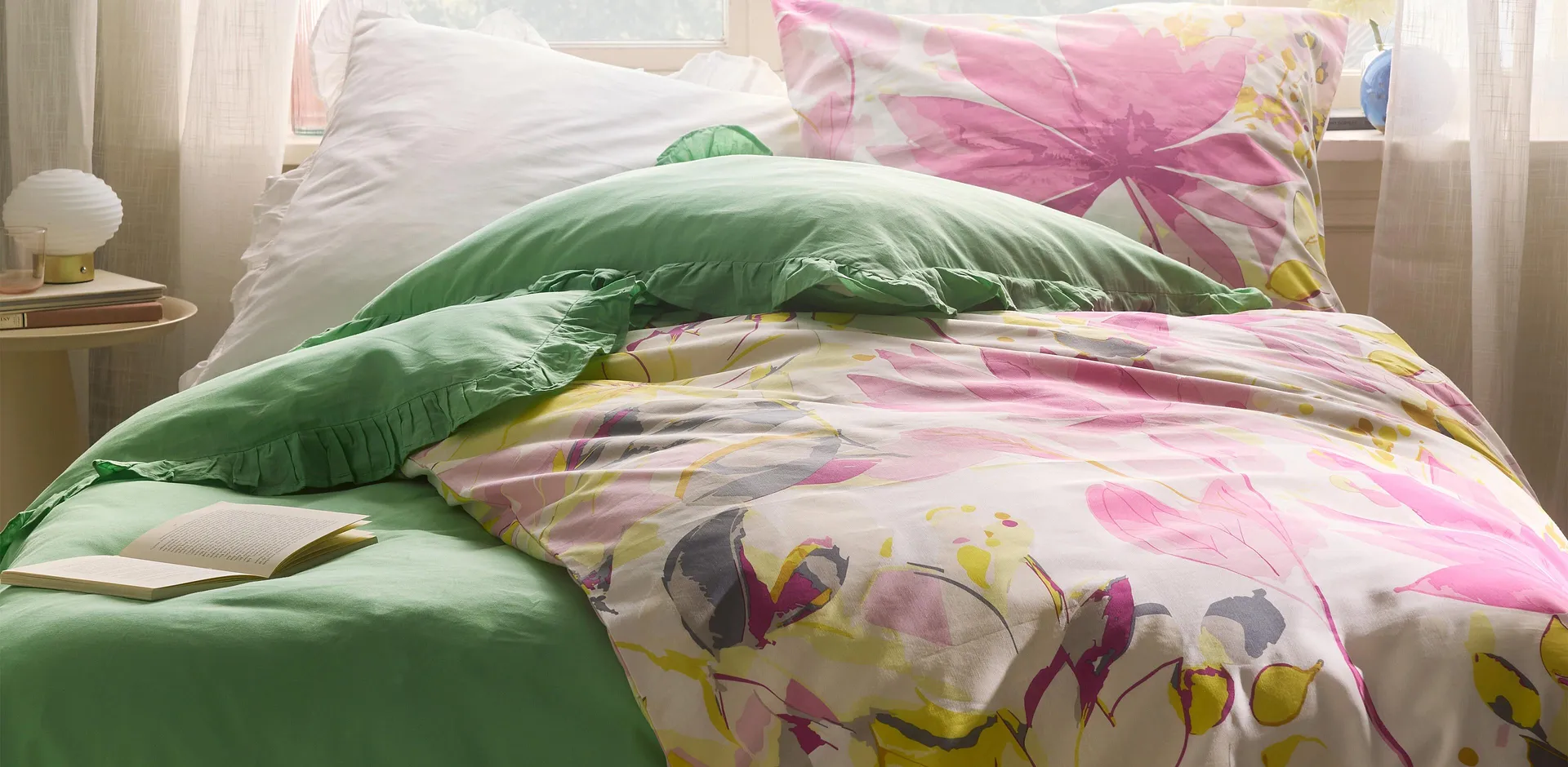 Farbenfrohe Bettw&auml;sche mit Blumenmuster, kombiniert mit gr&uuml;nem Kissen. Zarte Vorh&auml;nge rahmen das Bett ein.