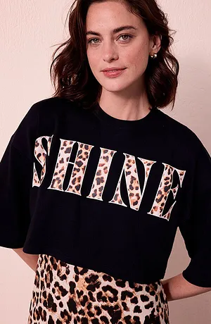 Femme portant un T-shirt avec SHINE en lettres léopard et une jupe léopard devant un mur clair