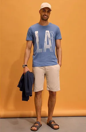 T-shirt bleu avec imprimé « LA », short clair, casquette blanche et sandales pour un look estival décontracté.