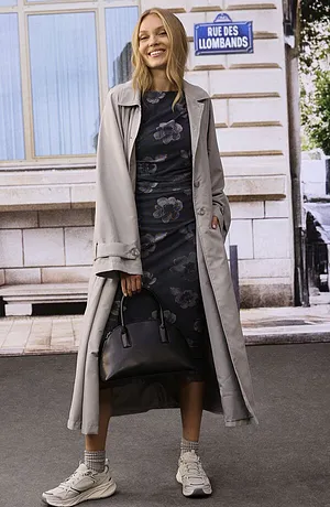 Trench-coat élégant porté sur une robe midi à fleurs, le tout complété par des baskets blanches et un sac à main noir.
