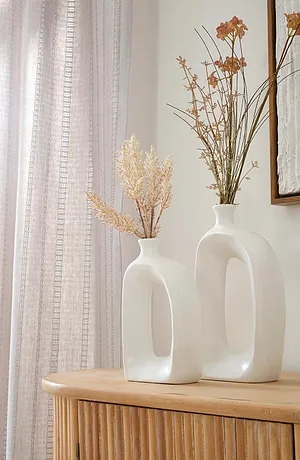 Des rideaux clairs et vaporeux encadrent la pièce, des vases blancs élégants garnis de fleurs séchées sont posés sur un meuble en bois.