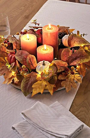 Herbstliche Tischdekoration mit Kerzen, umgeben von bunten Blättern auf weißem Tischtuch.