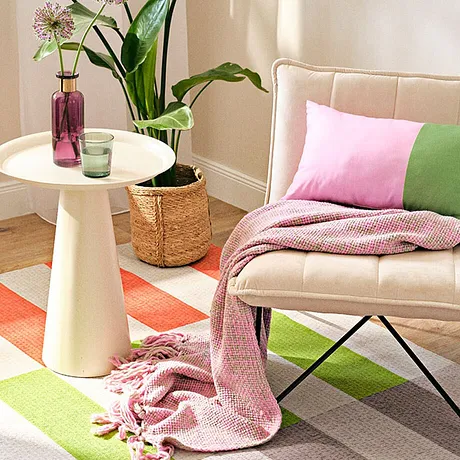 Textiles clairs dans des couleurs pastel : couverture rose, coussin vert et rose sur un fauteuil beige.