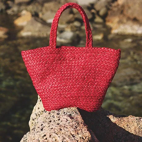 Leuchtend rote Strohhandtasche ruht auf Felsen am Wasser, perfekt für sommerliche Strandausflüge.