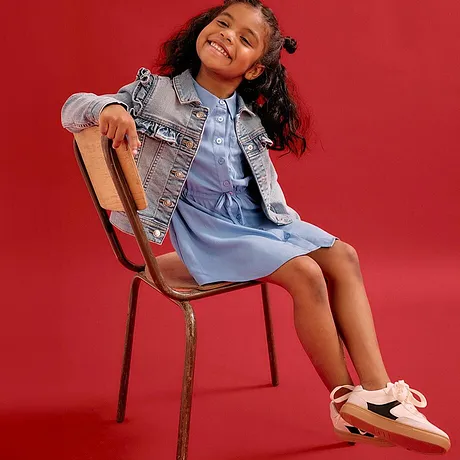 Petite fille joyeuse en robe bleu clair et veste en jean. Baskets confortables.