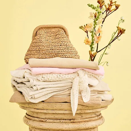 Tons naturels chaleureux : sac en paille, textiles raffinés et un vêtement blanc empilés sur une colonne en bois. 