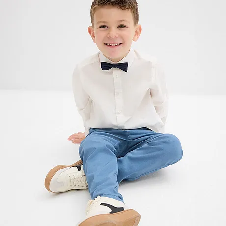 Eleganter Junge in weißem Hemd mit Fliege, hellblauer Hose und weißen Schuhen.