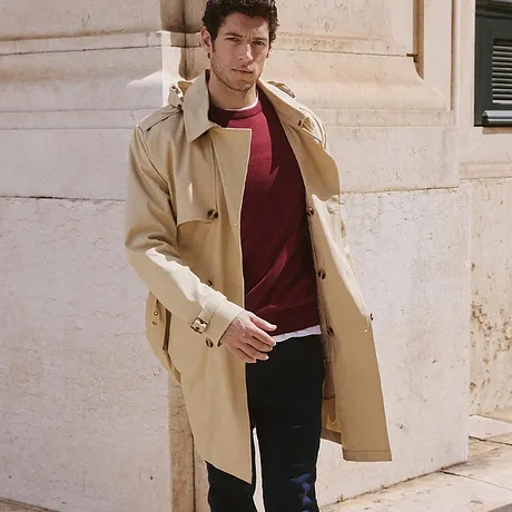 Eleganter beiger Trenchcoat über rotem Pullover und dunkler Jeans. 