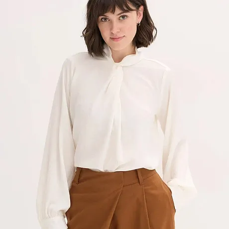 Blouse blanche à col noué, associée à un pantalon marron.