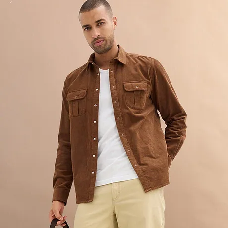 Chemise marron par dessus un T-shirt blanc, associée à un pantalon clair.