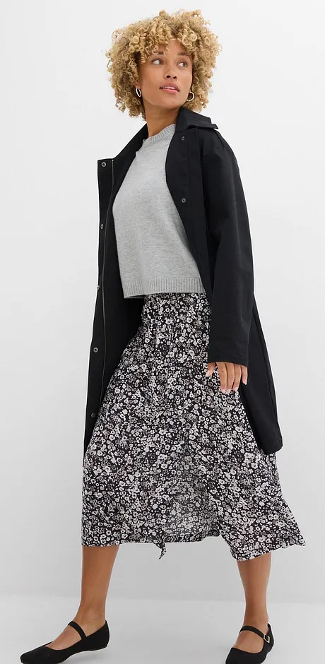 Eleganter schwarzer Mantel, graues Oberteil und langer gemusterter Rock kombiniert mit flachen Schuhen.