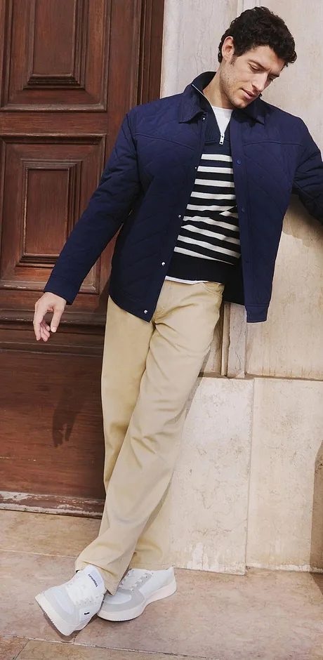 Eine dunkelblaue Jacke über einem gestreiften Oberteil, beige Hose und weiße Sneaker.