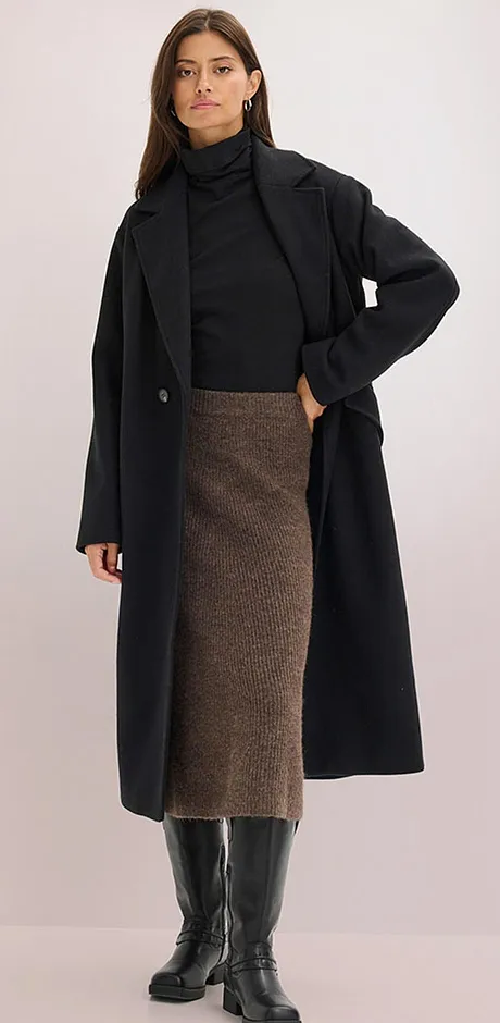 Manteau noir élégant sur un pull à col roulé, associé à une jupe midi marron et des bottes hautes.