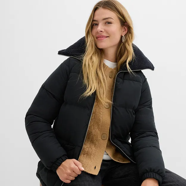 Schwarze Winterjacke mit hohem Kragen &uuml;ber gelbem Strickpullover und wei&szlig;em Shirt.