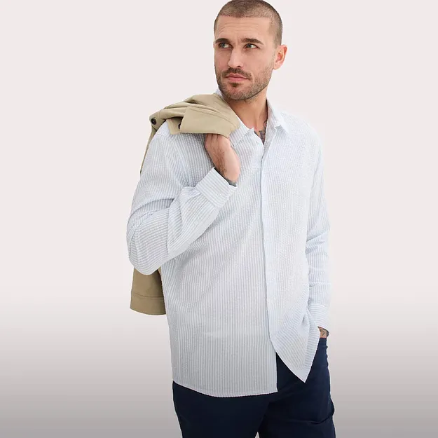 Chemise ray&eacute;e bleu clair, pantalon de couleur fonc&eacute;e et pull beige par dessus l'&eacute;paule.