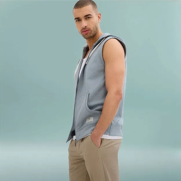 Mann tr&auml;gt hellblaue Sweatweste mit Kapuze &uuml;ber wei&szlig;em Shirt und beige Hose.