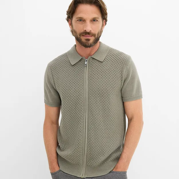Gr&uuml;nes Strickpolohemd mit Rei&szlig;verschluss und markantem Muster, kombiniert mit dunkler Jeans.