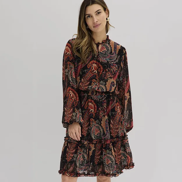 Sie tr&auml;gt ein flie&szlig;endes Kleid mit buntem Paisley-Muster und R&uuml;schendetails.