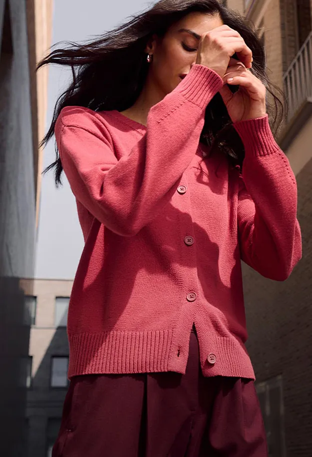 Sie tr&auml;gt eine leuchtend korallefarbene Strickjacke und eine weinrote Hose.