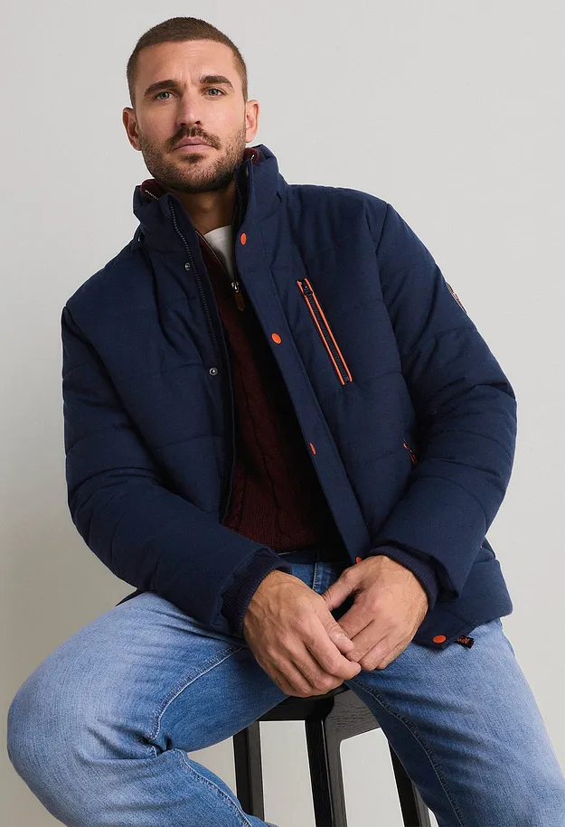 Dunkelblaue Steppjacke mit orangefarbenen Details, roter Strickpullover und helle Jeans.