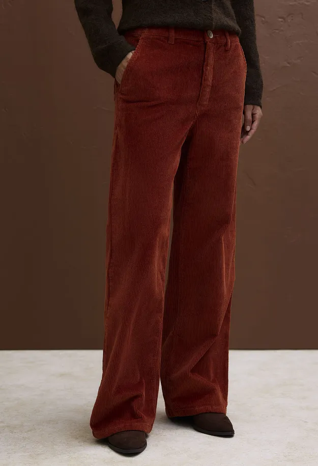 Sie tr&auml;gt eine gemusterte Strickjacke &uuml;ber einem wei&szlig;en Hemd und eine orange Cordhose.