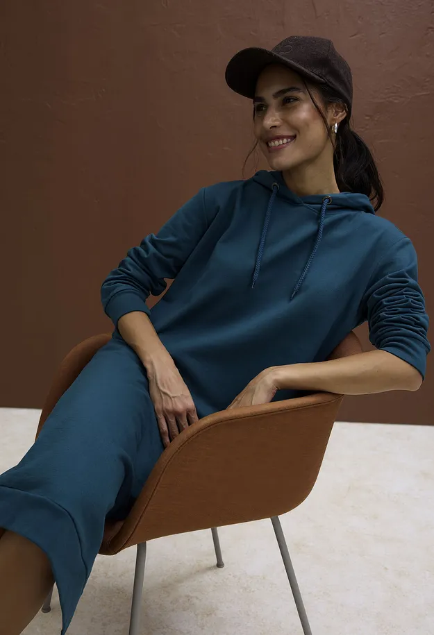 Sie tr&auml;gt ein t&uuml;rkisfarbenes Hoodie-Kleid und eine braune Baseballkappe.