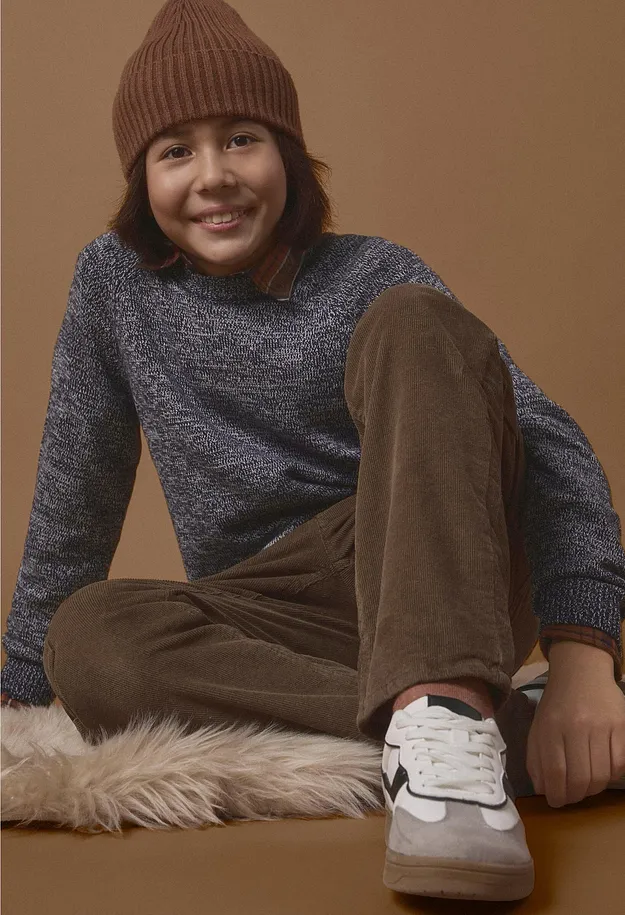 Ein M&auml;dchen tr&auml;gt eine braune Beanie, einen pinken Pullover, blaue Jeans und wei&szlig;e Sneaker.